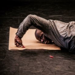 large black male in dark clothing lying face down on top of a piece of light brown butcher paper, that is on the floor. Only the upper half of the body is visible. the black male is using one arm to write on the paper with a red marker. The visible arm is bent at a slight right angle with the hand in a fisted position while writing. The marker's cap lies on the floor off of the paper.